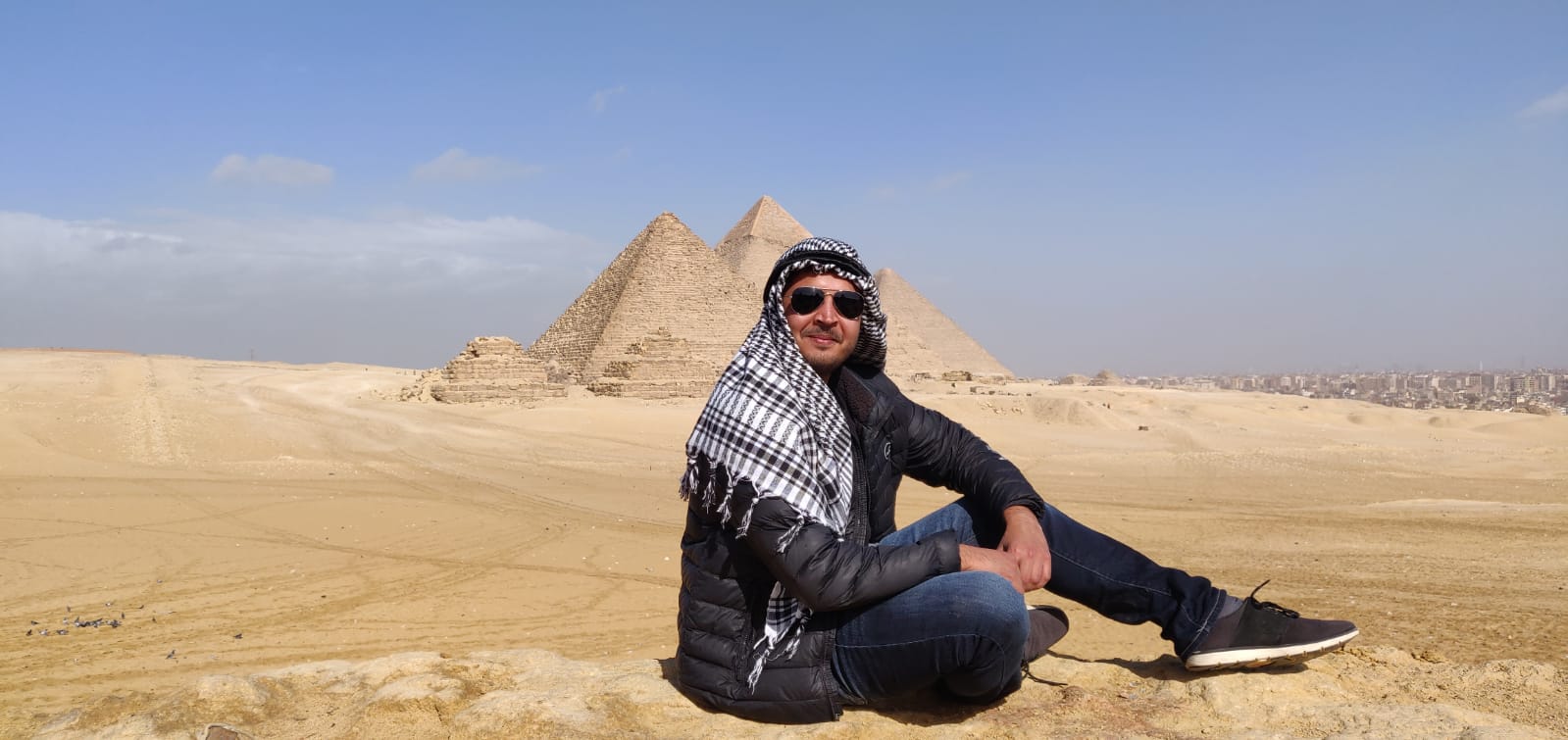 Founder in sunglasses and a checked headscarf seated on desert rock in front of the Pyramids of Giza